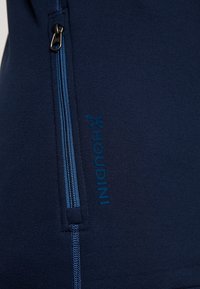 Tamnoplava flis tkanina s džepom na patentni zatvarač i plavim detaljem na zatvaraču. Sadrži logo u kontrastnoj plavoj boji, glatku teksturu i prilagođen oblik.