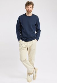 Sweatshirt marine avec des poignets et un col côtelés, arborant des accents aux épaules. Associé à un pantalon beige clair, décontracté, et des baskets crème.