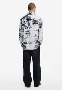 Long-sleeve shirt in white with black abstract patterns and text, featuring a relaxed fit and a straight hem, paired with wide black pants.
