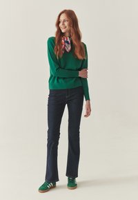 Vihreä neulepusero V-kaula-aukolla, tummat leveälahkeiset farkut ja vihreät tennarit valkoisilla raidoilla, yhdistettynä moniväriseen silkkihuiviin.