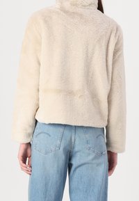 Faux fur jacket in beige with a high collar, cropped design, and long sleeves. Paired with mid-rise blue denim jeans featuring back pockets.