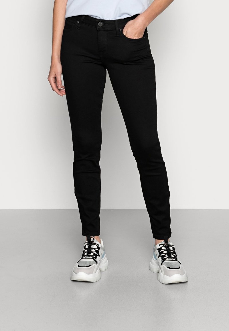 Jeans negros ajustados con una textura suave, que cuentan con bolsillos delanteros y un cierre de botón, combinados con zapatillas gruesas en blanco y gris.