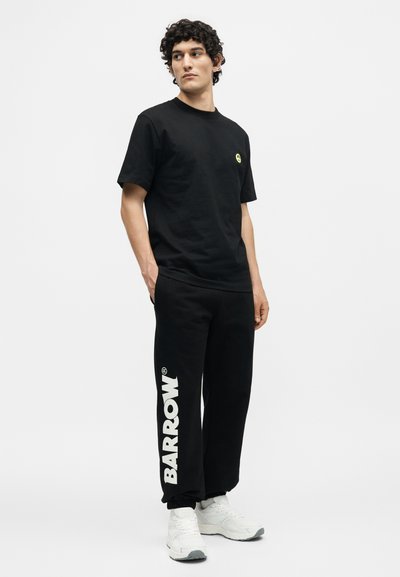 Νέος άνδρας που φοράει μαύρο μπλουζάκι και μαύρη φόρμα με το κείμενο «BARROW», στέκεται μπροστά σε ένα απλό λευκό φόντο.
