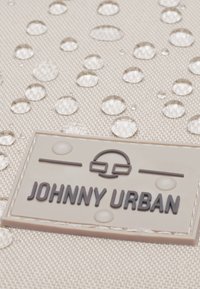 Beige textúrájú vászon vízcsepp manuálisan és egy téglalap alakú logó folt, amelyen sötét betűkkel olvasható a "JOHNNY URBAN" felirat.