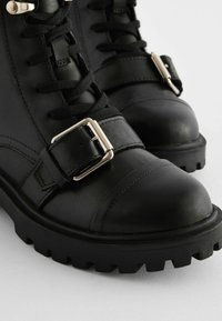 Svarta lädersneakers i ankellängd har en rund tå, snörning fram och ett band med en silverfärgad spänne. Sulan är texturerad med ett robust mönster.
