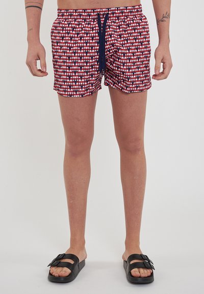 Costumi da bagno a righe rosse e bianche con un laccio nero, caratterizzati da una vita elastica e un tessuto leggero. Indossati con sandali neri.