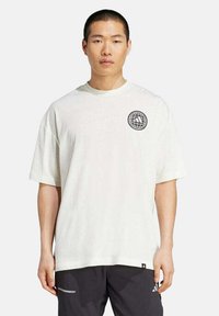 Mann mit einem lockeren weißen T-Shirt, das ein schwarzes Globus- und Streifenlogo auf der Brust hat, und einer schwarzen Shorts mit kleinen weißen Logos.