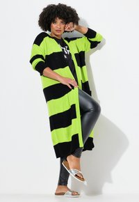 Cardigan lavorato a maglia a strisce verde brillante e nero, con una vestibilità ampia, polsini a coste e tasche laterali, abbinato a leggings neri e sandali argentati.