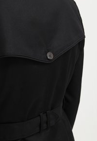 Abrigo negro tipo trench con cintura ceñida por un cinturón, gran solapa y detalles de botones en la solapa. El material parece suave con un acabado mate.