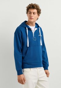 Junger Mann trägt blauen Kapuzenpullover mit weißen Kordeln und weißen Hosen, schaut leicht nach links vor neutralem Hintergrund.