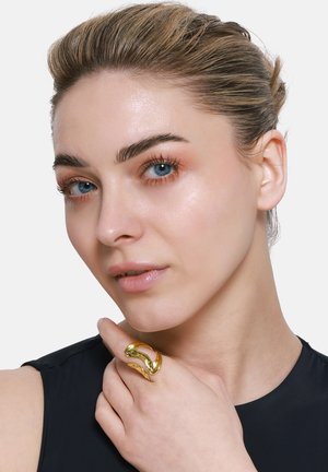Goldring mit gewelltem Design, das glatte, polierte Oberflächen und eine offene Aussparung aufweist. Modell trägt es zu einem dunklen Oberteil.