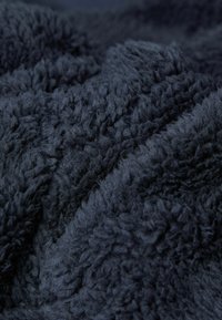 Tmavě modrá, sametová tkanina s nadýchanou strukturou a hustým vlasem, ukazující nepravidelné vzory a měkké, silné vlákna uspořádaná ve skládání.