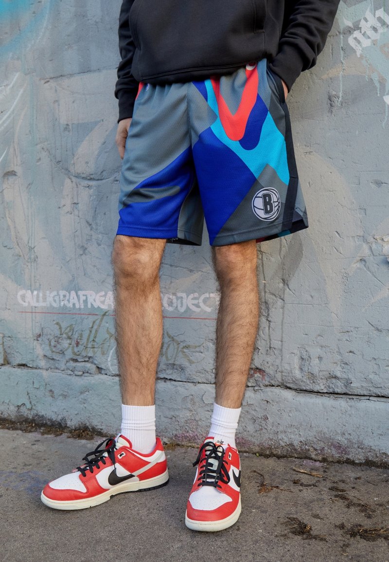 Basketball shorts in gray with blue, red, and black geometric patterns; elastic waistband; logo on left leg. Worn with red and white sneakers.