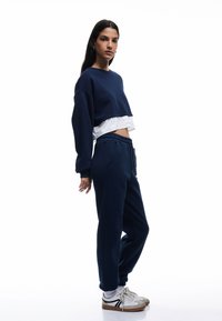 Marineblå cropped sweatshirt med et hvidt lag under, parret med marineblå sweatpants og hvide sneakers med brune accenter. Glat tekstur.