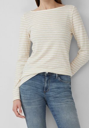 Femme portant un haut à manches longues rayé crème et beige, associé à un jean bleu en denim, debout devant un fond uni gris clair.