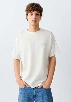 Jonge man met krullend haar, een wit T-shirt met een klein logo op de borst en blauwe jeans, handen in de zakken, neutrale achtergrond.