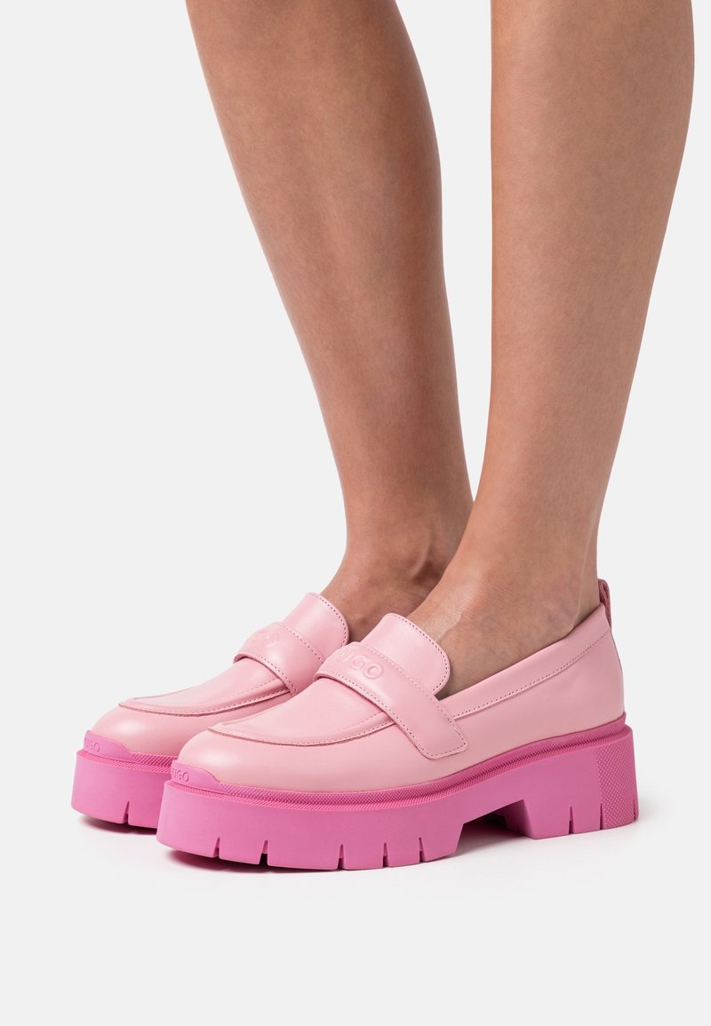 Personne portant des mocassins en cuir rose clair avec des semelles épaisses et volumineuses de couleur rose vif, debout devant un fond clair uni.
