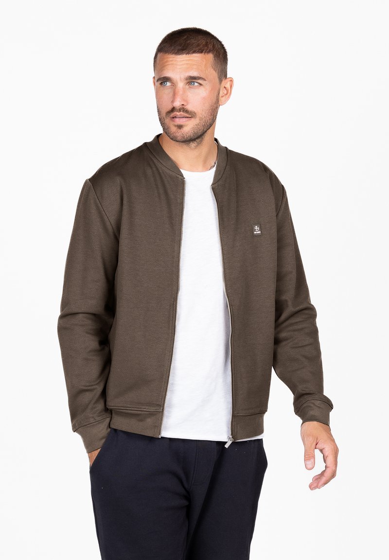 Chaqueta bomber de color marrón oscuro con cremallera, puños y cuello acanalados, hecha de un tejido texturizado, que presenta un pequeño logo en el lado izquierdo del pecho. Usada sobre una camiseta blanca.