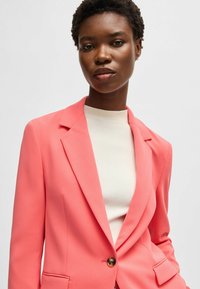 Korallfärgad kavaj med slag och en enkel knappstängning, kombinerad med en krämfärgad ribbad topp. Slät tyg, skräddarsydd passform, diskreta fickor.