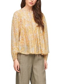 Blusa gialla a fantasia con texture plissettata e maniche lunghe, caratterizzata da disegni floreali in diverse tonalità. Indossata su pantaloni beige larghi.