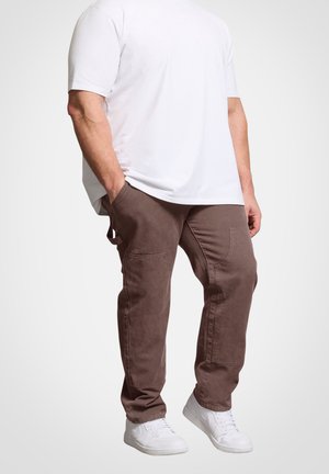 Mand iført en almindelig hvid kortærmet skjorte, brune cargobukser med sidelommer og hvide sneakers, står med hænderne i lommerne.