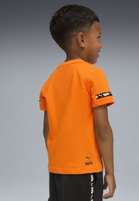 Junge, der nach rechts schaut, trägt ein leuchtend oranges T-Shirt mit kurzen Ärmeln und schwarzem Logo auf dem Arm und dem Saum sowie schwarze Hosen mit weißen Logos an der Seite.
