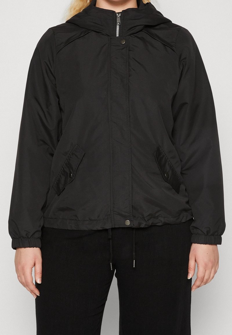 Persona con chaqueta negra con capucha y cremallera, bolsillos con botones a presión y puños elásticos, de pie frente a un fondo gris liso.