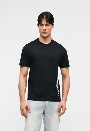 Meesmodell kannab musta lühikese varrukaga T-särki, millel on valged vertikaalsed tähed küljel, kombineeritud heleroheliste teksadega, seistes halli tausta ees.