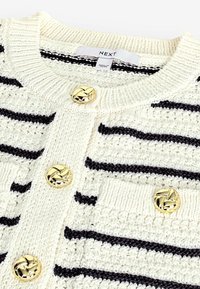 Cardigan crema con righe nere, texture a maglia e bottoni intrecciati dorati. Presenta due tasche frontali con colletto a coste.