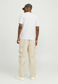 Camiseta blanca de manga corta, pantalones cargo beige con múltiples bolsillos y zapatillas blancas. Gorra negra llevada en un ángulo. Vista trasera.