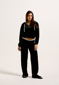 Conjunto de loungewear em veludo negro, composto por um hoodie cropped com fecho e calças largas, destacando-se uma cintura com logótipo branco. Textura suave.