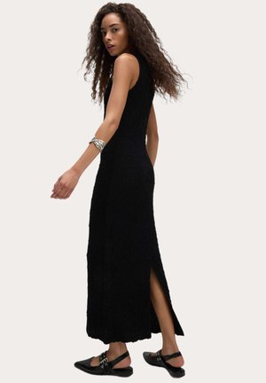Abito nero senza maniche con un design testurizzato, fessure laterali per la mobilità e una silhouette lunga e aderente. Abbinato a scarpe nere piatte.