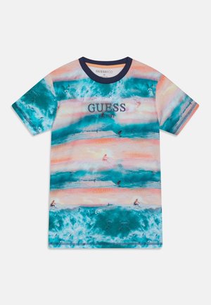 T-shirt met korte mouwen met een patroon van blauwe golven en strand, met de tekst "GUESS est. 1981" op de borst en een donkerblauwe kraag.