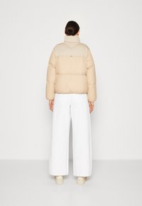 Béžová péřová bunda s vysokým límcem a staženým pasem. Zadní strana má barevně členěný design, doplněný o bílé kalhoty s širokými nohavicemi.