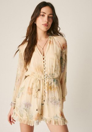 Tânără cu păr lung și castaniu, purtând o rochie ușoară cu model floral, nasturi, o curea în talie și tiv cu volane, pe un fundal neutru.