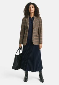 Brown check blazer over a navy polo, paired with a pleated navy skirt. Black leather tote bag and knee-high black boots complete the look.