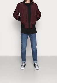 Chaqueta bomber burdeos con forro acolchado, puños y dobladillo acanalados; combinada con una camiseta negra de gran tamaño y jeans de mezclilla azules. Zapatillas altas negras.