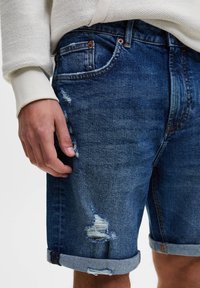 Pantaloncini in denim di colore blu scuro con dettagli strappati e orlo arrotolato. Presentano tasche anteriori e una chiusura standard con bottone e zip.