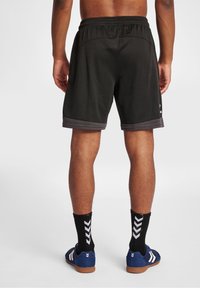 Pantalones cortos atléticos negros con un borde gris en el dobladillo, que cuentan con una cinturilla ajustable y una textura suave. Se llevan con zapatillas de deporte azules y calcetines negros.