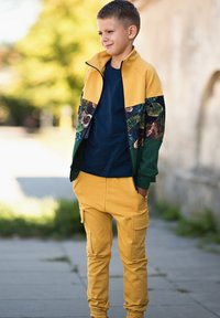 Dziecięcy dres z żółtą, zapinaną na zamek kurtką z wzorzystymi rękawami i jednokolorowymi zielonymi akcentami, w zestawie z żółtymi spodniami bojówkami.