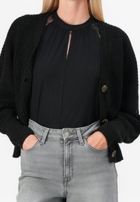 Cárdigan negro de punto con cierre de botones, con puños acanalados. Debajo, una blusa negra con diseño de ojal. Jeans gris de cintura alta.