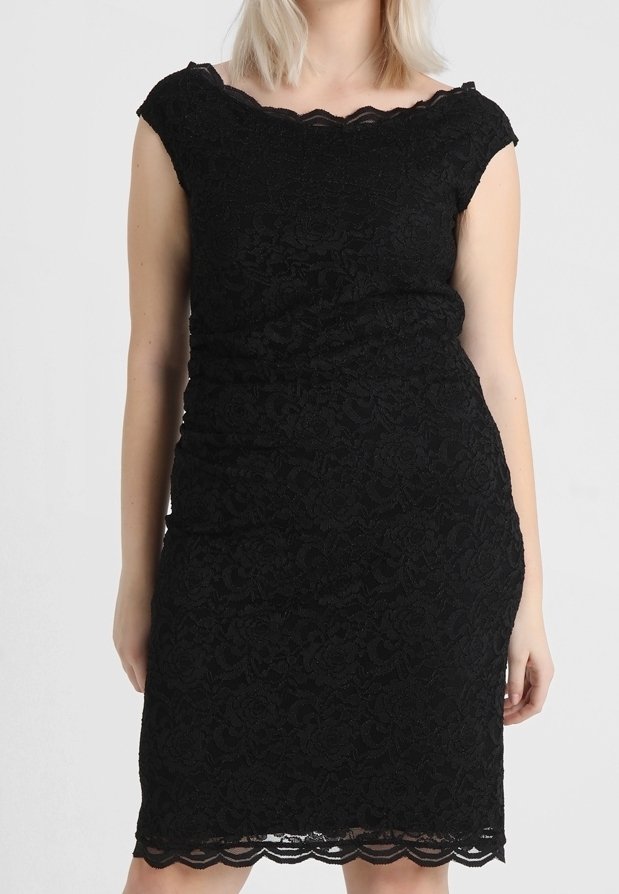 Femme portant une robe en dentelle noire ajustée avec des manches courtes et un ourlet festonné, debout devant un fond clair uni.