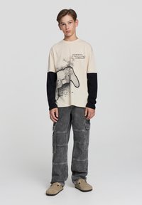 T-shirt beige con maniche lunghe nere, con grafica di un controller di gioco. Pantaloni cargo grigi con più tasche e una vestibilità rilassata.