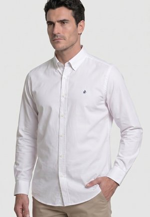 Hombre con camisa blanca de manga larga abotonada con cuello, pantalones beige, de pie con una mano en el bolsillo contra un fondo claro y liso.