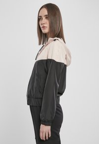 Svart og blush vindjakke med hette, elastiske mansjetter og nederkant; har sidelommer og en glatt, blank overflate.