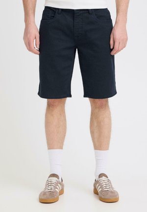 Mand iført sorte denimshorts, hvide crew-sokker og beige ruskindssneakers, stående mod en ensfarvet baggrund.
