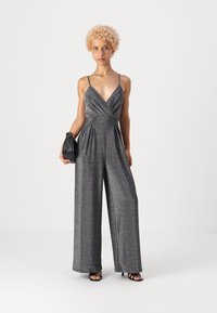 Γυαλιστερή, μεταλλική γκρίζα φόρμα με βαθύ V-λαιμό, λεπτές τιράντες και φαρδιές γάμπες. Συνδυασμένη με μια μικρή μαύρη τσάντα ώμου και λουριά παπούτσια.