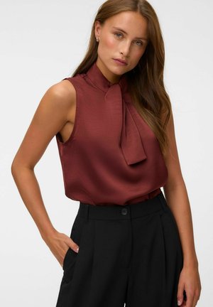 Femme aux longs cheveux bruns portant un chemisier bordeaux sans manches avec un détail nœud papillon et un pantalon noir taille haute, main dans la poche.