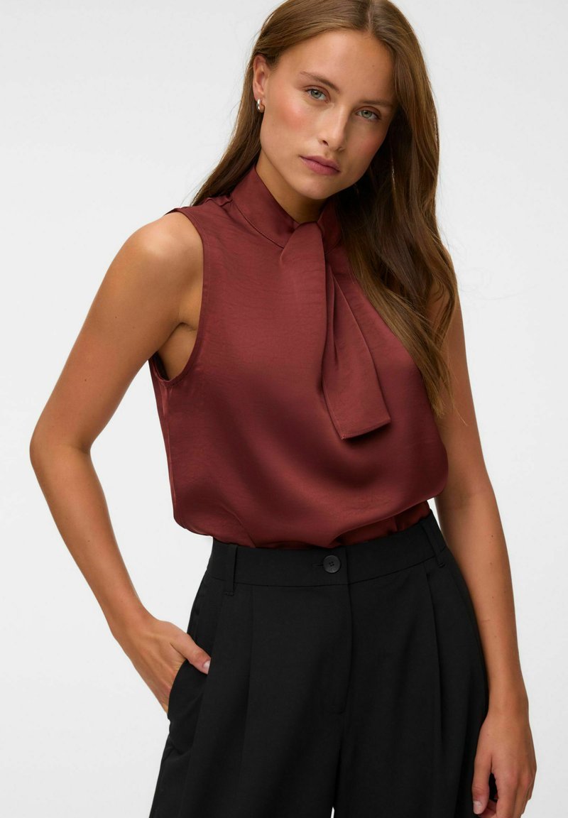 Femme aux longs cheveux bruns portant un chemisier bordeaux sans manches avec un détail nœud papillon et un pantalon noir taille haute, main dans la poche.