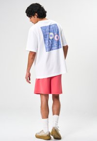 Weißes Baumwoll-T-Shirt mit einem blauen Gitterdesign und zwei runden Akzenten auf dem Rücken. Kombiniert mit korallenfarbenen Shorts und hellen Turnschuhen.